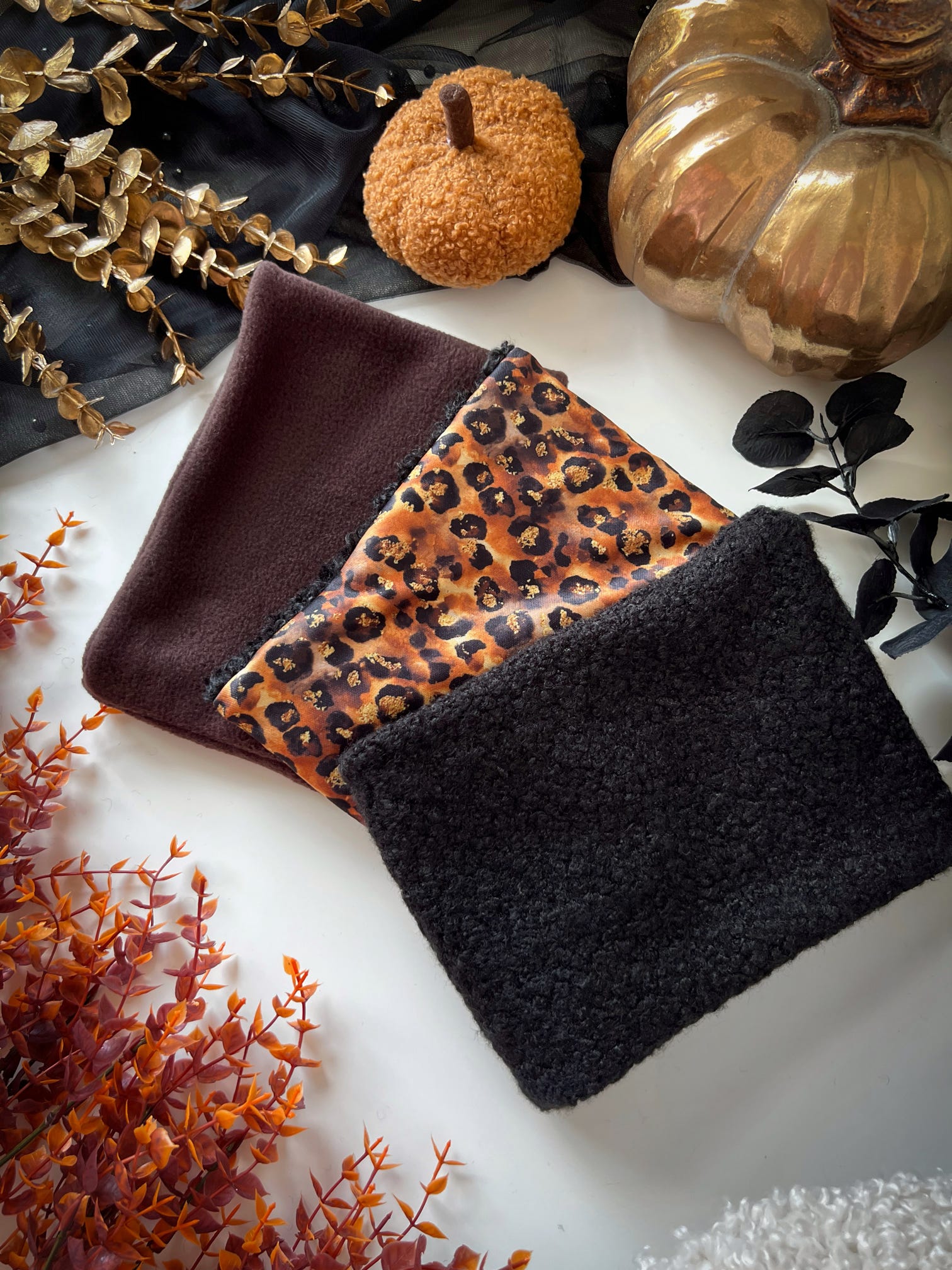 Three dog snoods in brown, leopard print, and black on a decorative surface with pumpkins and leaves.