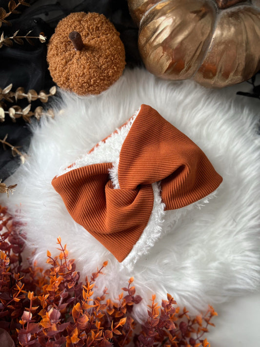 Orange knitted dog snood on a white fluffy surface with pumpkins and dried foliage.