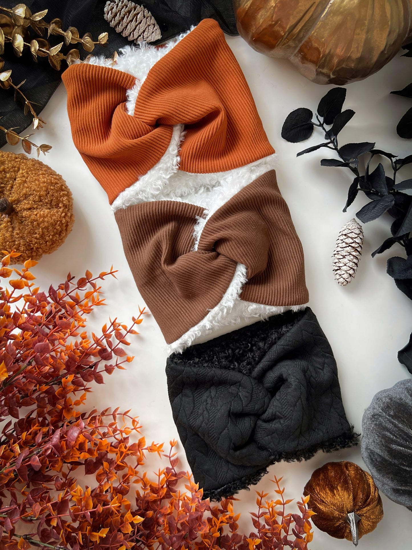 Three fabric dog snoods in orange, brown, and black on a white surface with autumn-themed decorations.